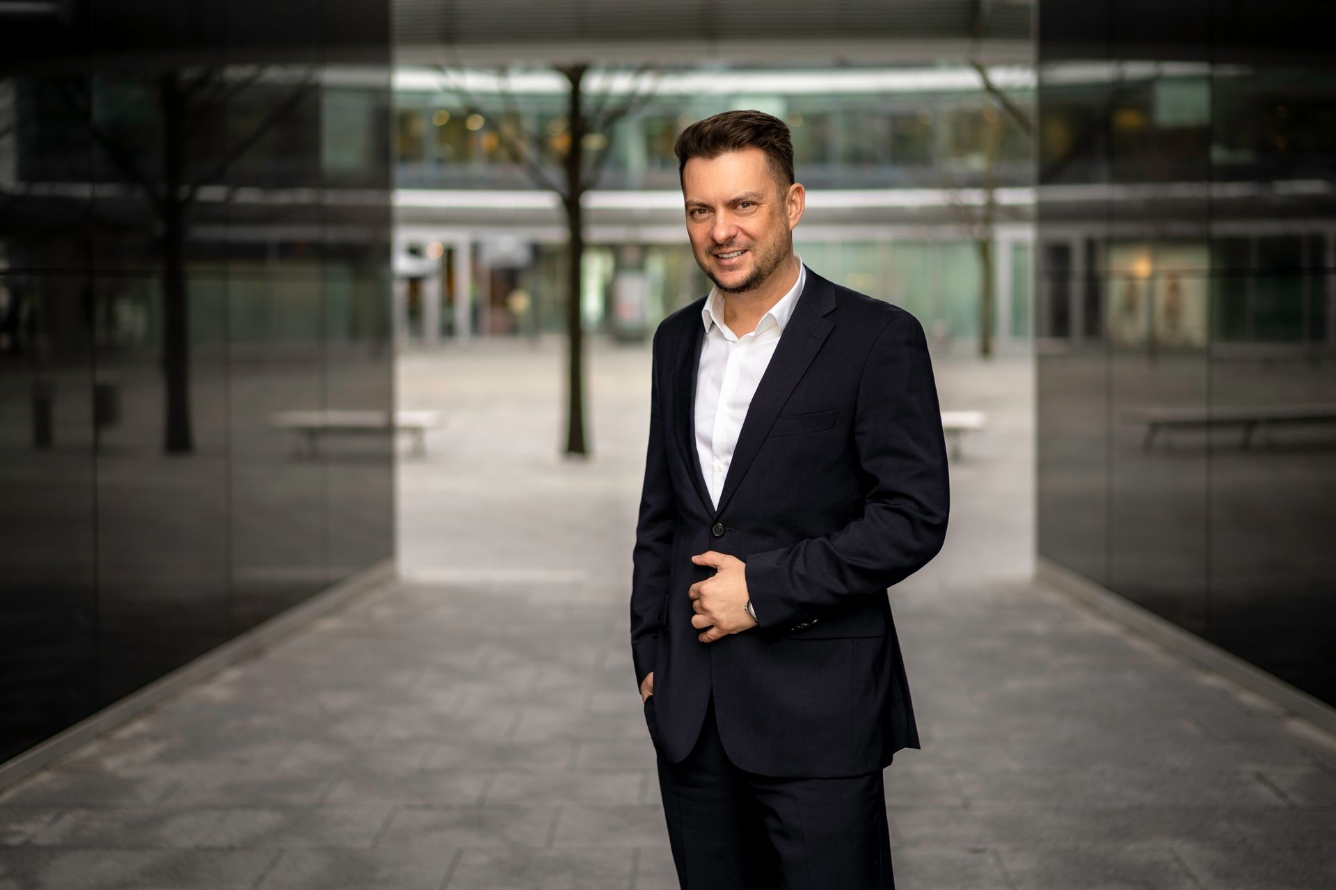 Man in a black suit standing outdoors in an urban setting with glass walls and trees.